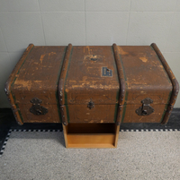 Auf einem Hocker vor einer Wand steht ein großer, alter Koffer mit deutlichen Gebrauchsspuren.