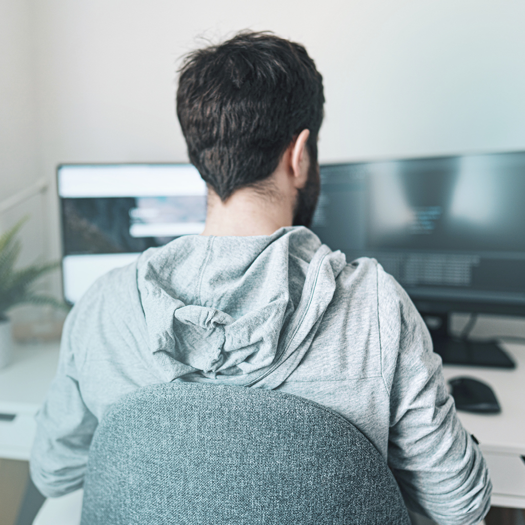Ein Mann in einem grauen Pullover sitzt an einem Schreibtisch, mit mehreren Bildschirmen, und arbeitet.