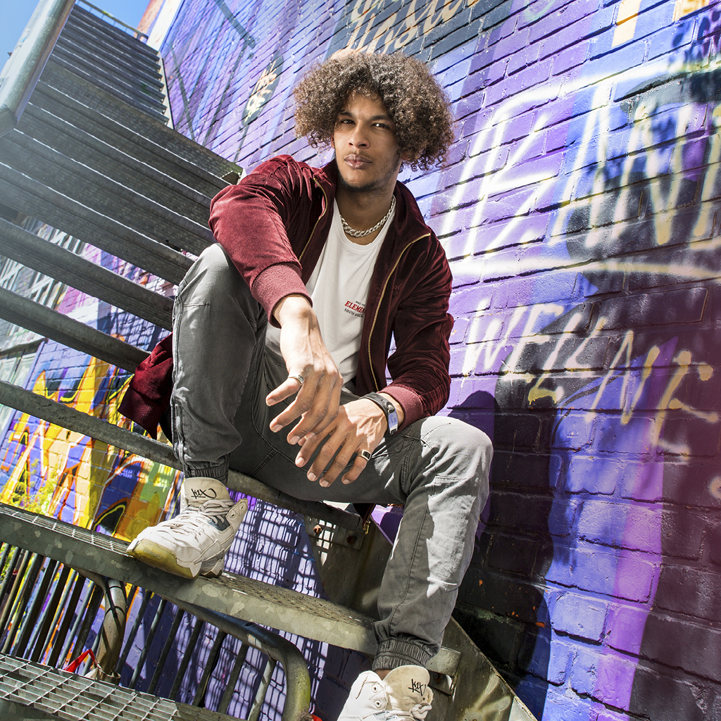 Ein junger Mann sitzt auf der Treppe vor einer Graffiti-Wand und post cool in die Kamera.