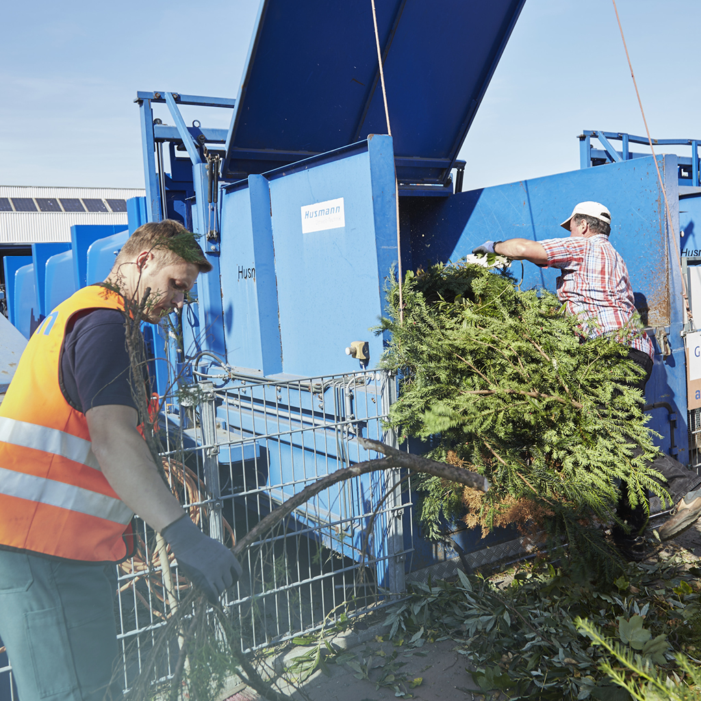 Ein Mann entsorgt Zweige und Äste in einem dafür vorgesehenen, blauen Container auf einem Recyclinghof, ein Mitarbeiter dessen trägt eine Warmweste und hilft ihm bei der richtigen Entsorgung.