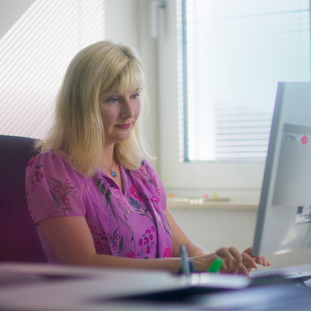 Eine blonde Frau mit einer pinken Bluse sitzt vor einem Computer und arbeitet daran.