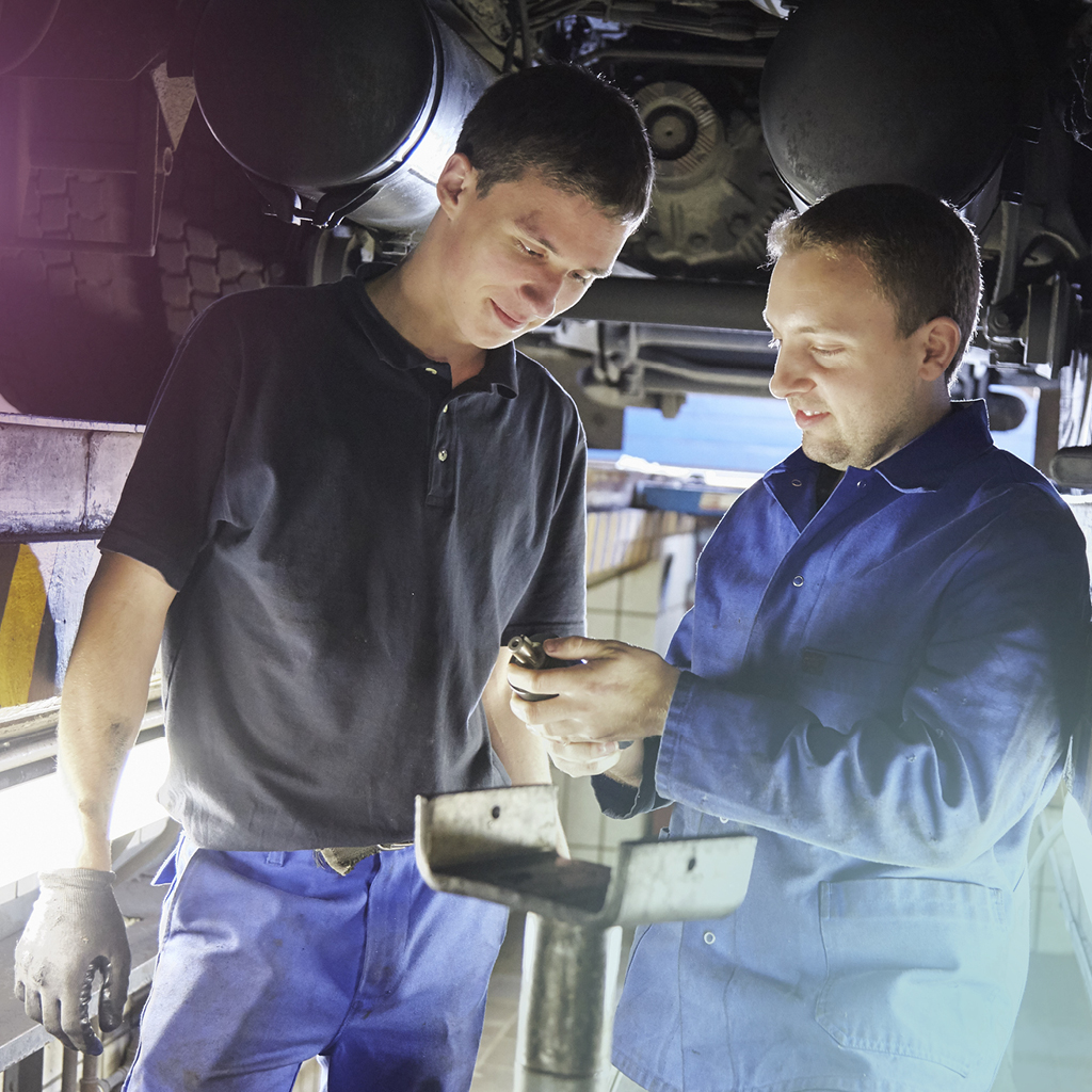 Zwei Techniker stehen in einer Werkstatt und reparieren gemeinsam etwas an einem Teil eines Wagens.