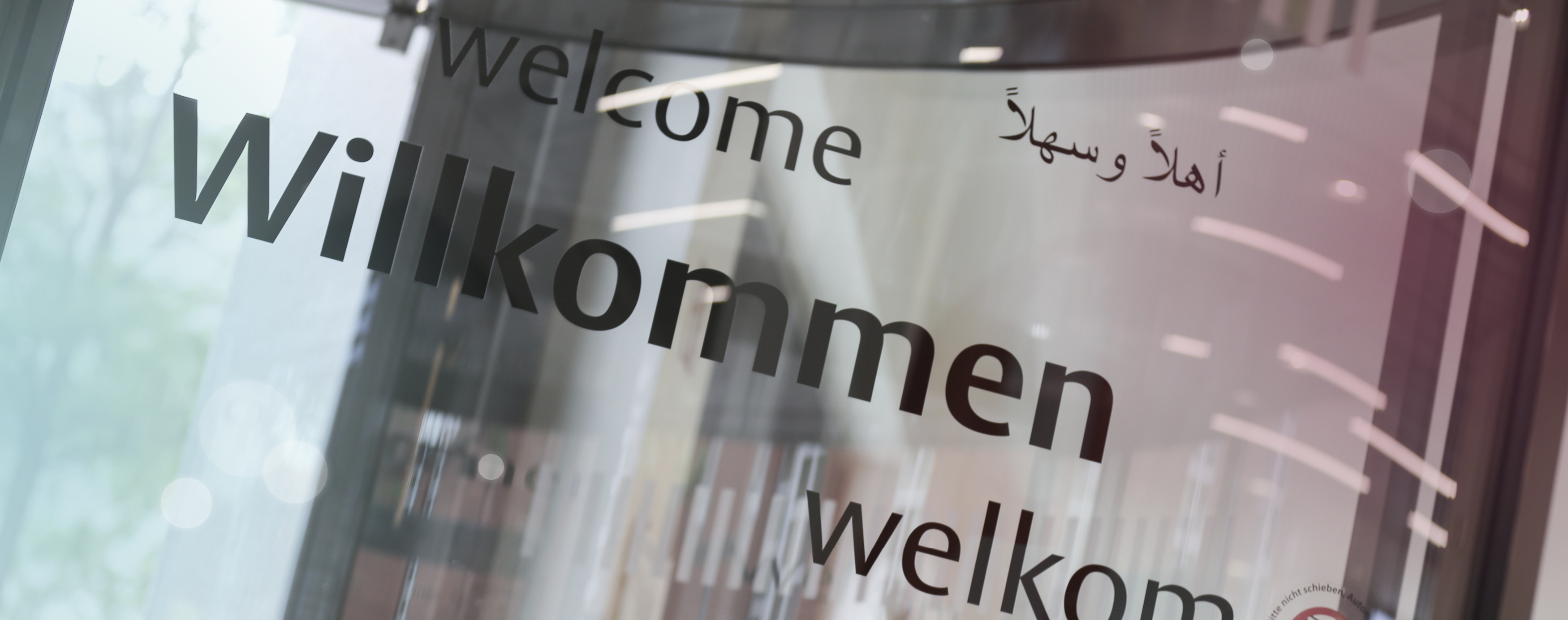 Die Glastür am Eingang des Servicezentrums Münster zeigt durch die Beschriftungen auf verschiedenen Sprachen, dass jeder bei der Stadt willkommen ist.