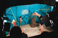 Zu sehen ist eine kleine Puppenbühne mit blauem Hintergrund auf dem steht 'Was können wir tun?'. Auf der kleinen Bühne steht ein Baum in Spielzeuggröße und drei Barbiefiguren, die von Händen gehalten werden. Im Vordergrund ist der Scheinwerfer einer Kamera zu sehen.