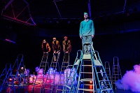Auf der Bühne sind viele hohe Trittleitern zu sehen. Auf vier Leitern in der Höhe stehen vier Akteure und blicken ins Publikum. Im blau-rosa angestrahlten Nebel sitzt links ein Schlagzeuger an seinem Instrument und spielt.