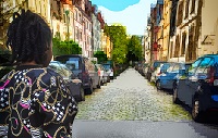Zu sehen ist eine Straße mit Autos und Häusern zu beiden Seiten, die mit einem Computerprogramm bearbeitet wurden. Links im Vordergrund steht eine Jugendliche. Sie schaut in die Straße.