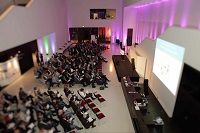 Bild von Veranstaltung mit Saalbestuhlung im großzügigen Foyer des LWL-Museums für Kunst und Kultur in Münster im Jahr 2017. Der Raum liegt im Halbdunkel, vor dem Treppenaufgang rechts im Bild ist eine Bühne aufgebaut, die weißen Wände sind teilweise violett angestrahlt.
