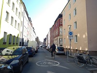 Blick auf die Fahrradstraße - rundes Piktogramm Fahrrad auf der Straße - rechts das Schild "Fahrradstraße. 2 Fahrradfahrer befahren die Straße, rechts 2 Fahrradbügel. Links und rechts der Straße parkende Autos, im Anschluss daran der Fußweg und die angrenzende Mehrfamilienhausbebauung.