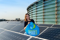 Jana Lange sitzt auf einem Dach mit Photovoltaikanlage und präsentiert das Logo der Stadt Münster: "Unser Klima 2030"