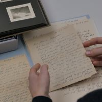 Eine Frau begutachtet einen alten Feldpostbrief in Sütterlin, im Hintergrund sind weitere Briefe und ein Fotoalbum zu sehen.