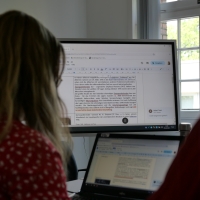 Im Vordergrund sitzen zwei Personen, die auf einen Laptopbildschirm und dahinter einen größeren Touchmonitor blicken, auf dem Texte projiziert werden, an denen sie per Textprogramm arbeiten.