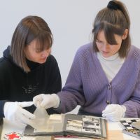 Two employees bend over an old photo album with black and white pictures and leaf through them. On the table are other items from the collection, such as an armband with the symbol of the Red Cross, various books and a board game.