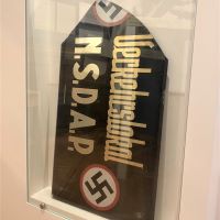 Ein schwarzes Schild steht in einer Vitrine. Darauf steht in Gold der Schriftzug 'Verkehrslokal NSDAP'. Auf beiden Seiten des Schriftzugs stehen Hakenkreuze, wobei das Hakenkreuz an der linken Seite nicht mehr vollständig ist, weil Teile des Schilds abgebrochen wurden.