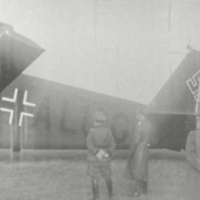 Drei Männer stehen auf einem Feld mit dem Rücken zur Kamera vor einem Flugzeug, dessen hintere Hälfte zu sehen ist.