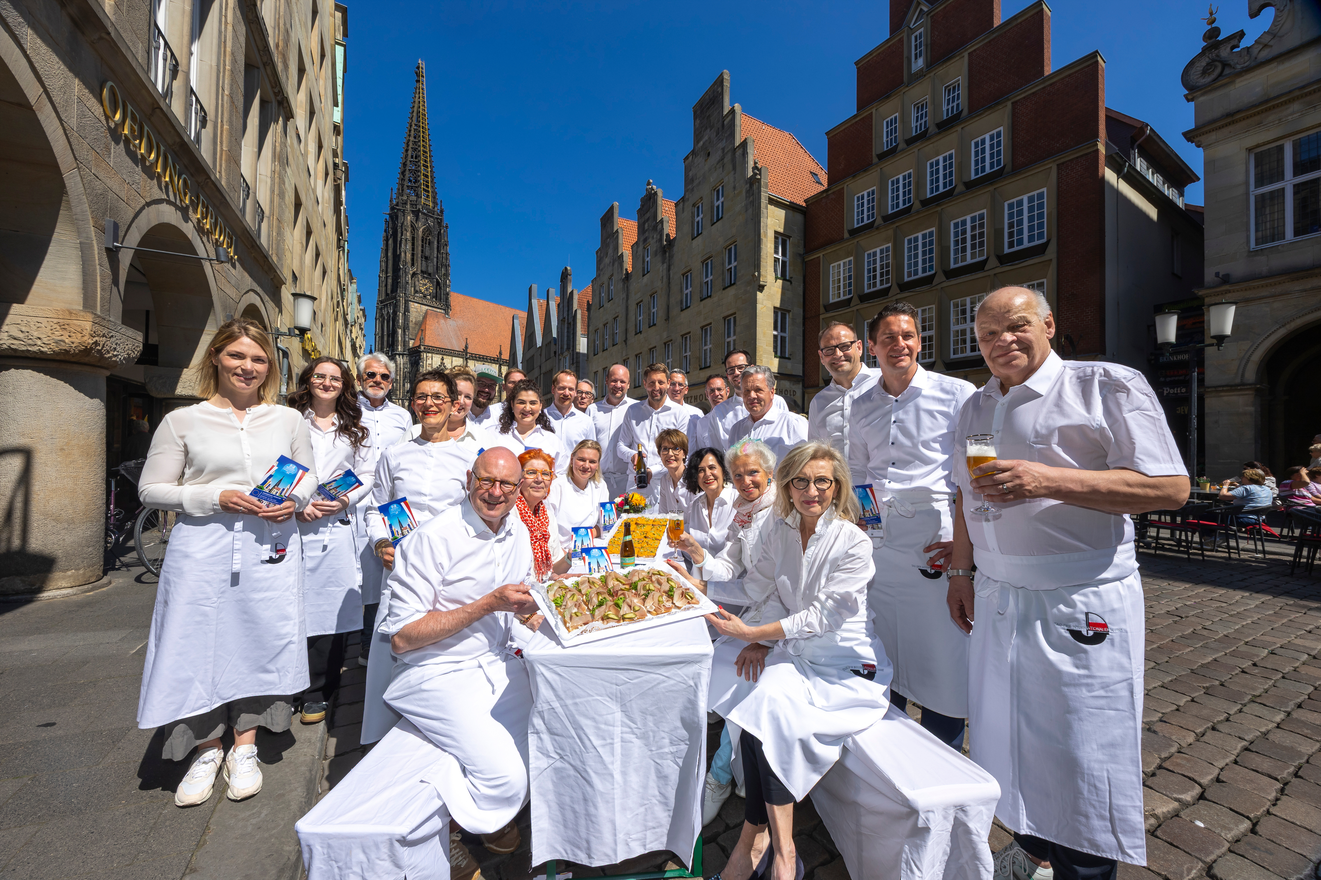 Weiß gekleidete Menschen sitzen an einem Tisch mit weißem Tischtuch oder stehen dahinter und präsentieren Schnittchen auf dem Prinzipalmarkt mit der Lambertikirche im Hintergrund