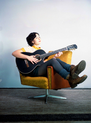 Die Liedermacherin Paula Linke sitzt mit einer Gitarre quer auf einem Sessel. Ihre Beine befinden sich über der Armlehne.