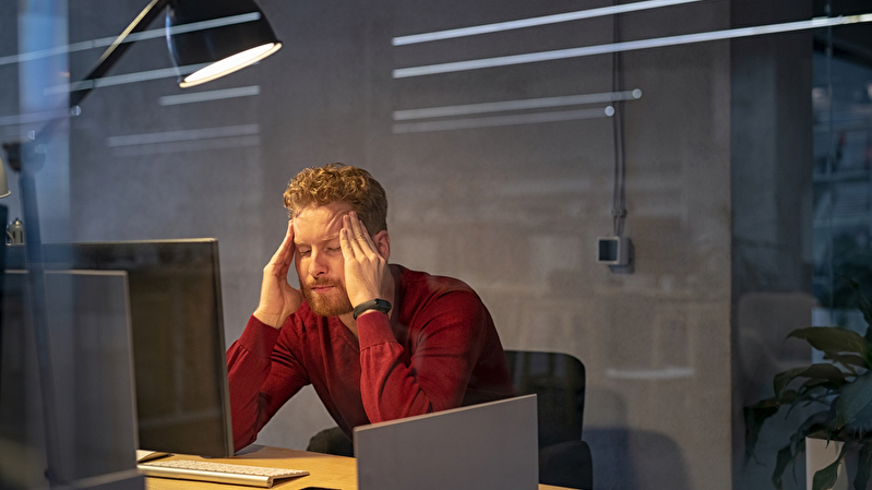 Ein junger Mann sitzt überarbeitet am Schreibtisch und massiert sich die Schläfen.
