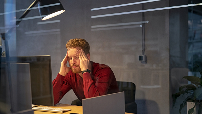 Ein junger Mann sitzt überarbeitet am Schreibtisch und massiert sich die Schläfen.