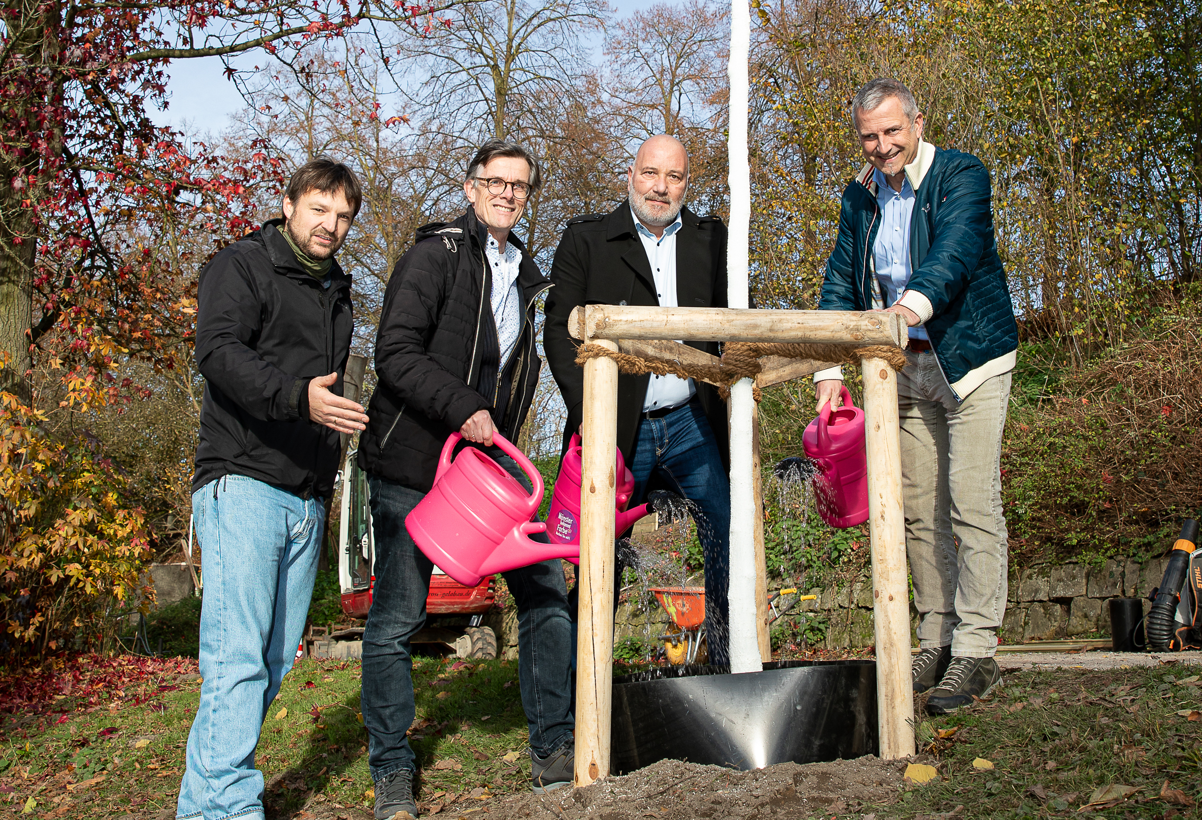 Vier Männer gruppieren sich um einen frisch geplanzten Baum, einer trägt eine Gießkanne.