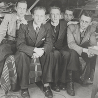 Ein Schwarzweißfoto von fünf jungen Männern. Sie tragen Anzüge und Lederschuhe, zwei von ihnen lächeln in die Kamera. Sie sitzen eng nebeneinander in einem mit Papier vollgestellten Raum.