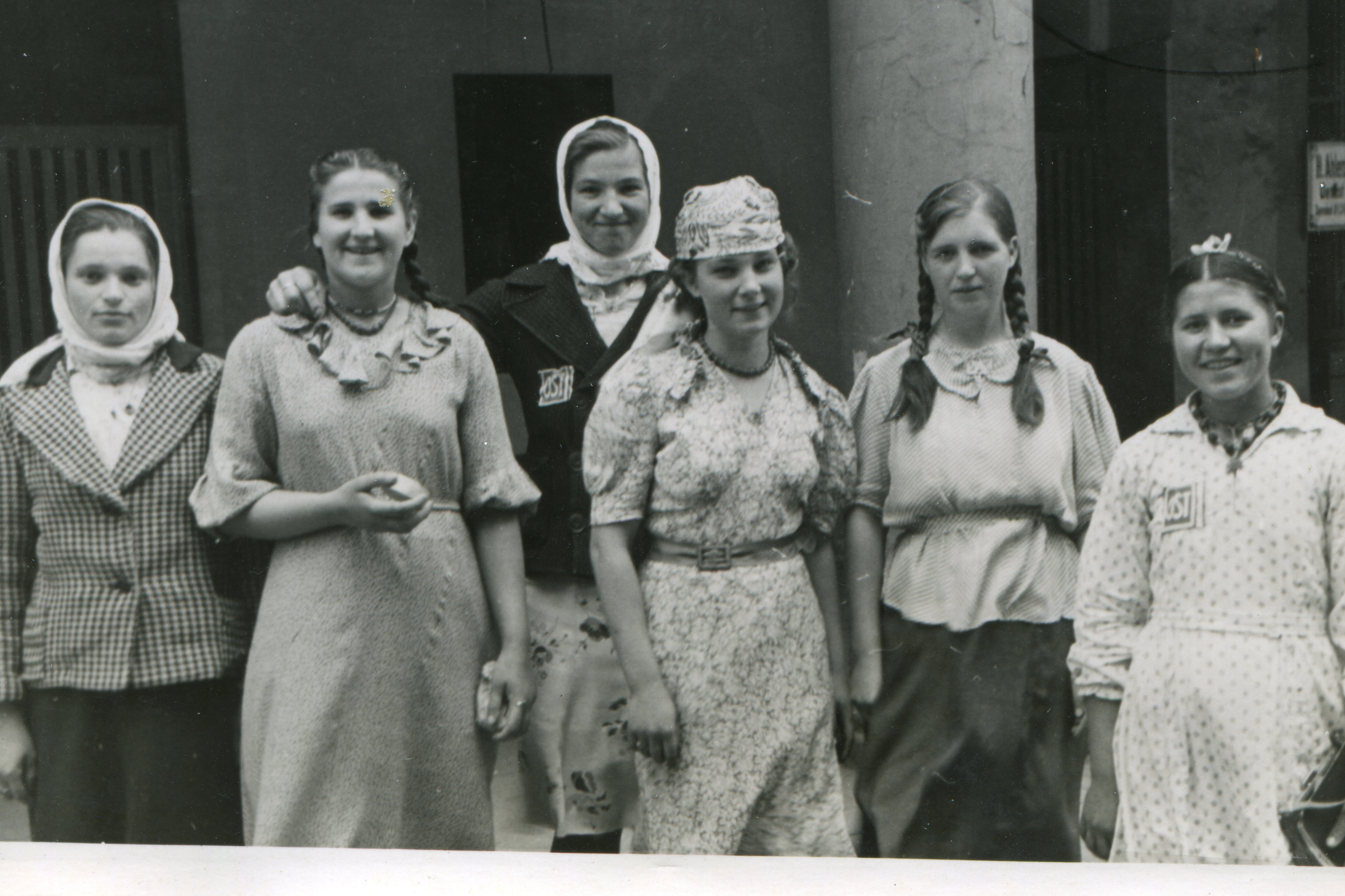 Sechs junge Frauen stehen nebeneinander und lächeln in die Kamera. Zwei von ihnen tragen auf der Brust das sogenannte „Ost“-Kennzeichen.