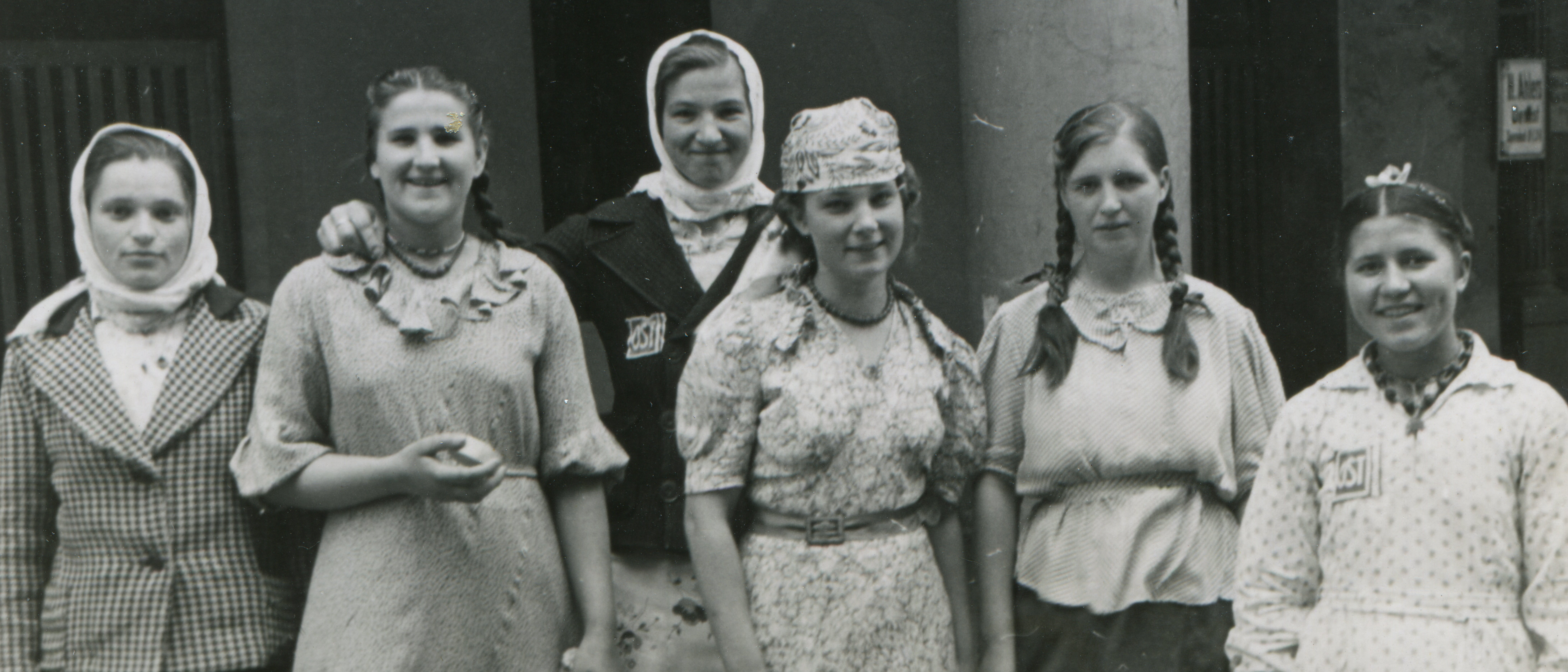 Sechs junge Frauen stehen nebeneinander und lächeln in die Kamera. Zwei von ihnen tragen auf der Brust das sogenannte „Ost“-Kennzeichen.