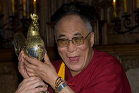 Der Dalai Lama in rot und gelb gekleidet hält den Goldenen Hahn in den Händen und freut sich.