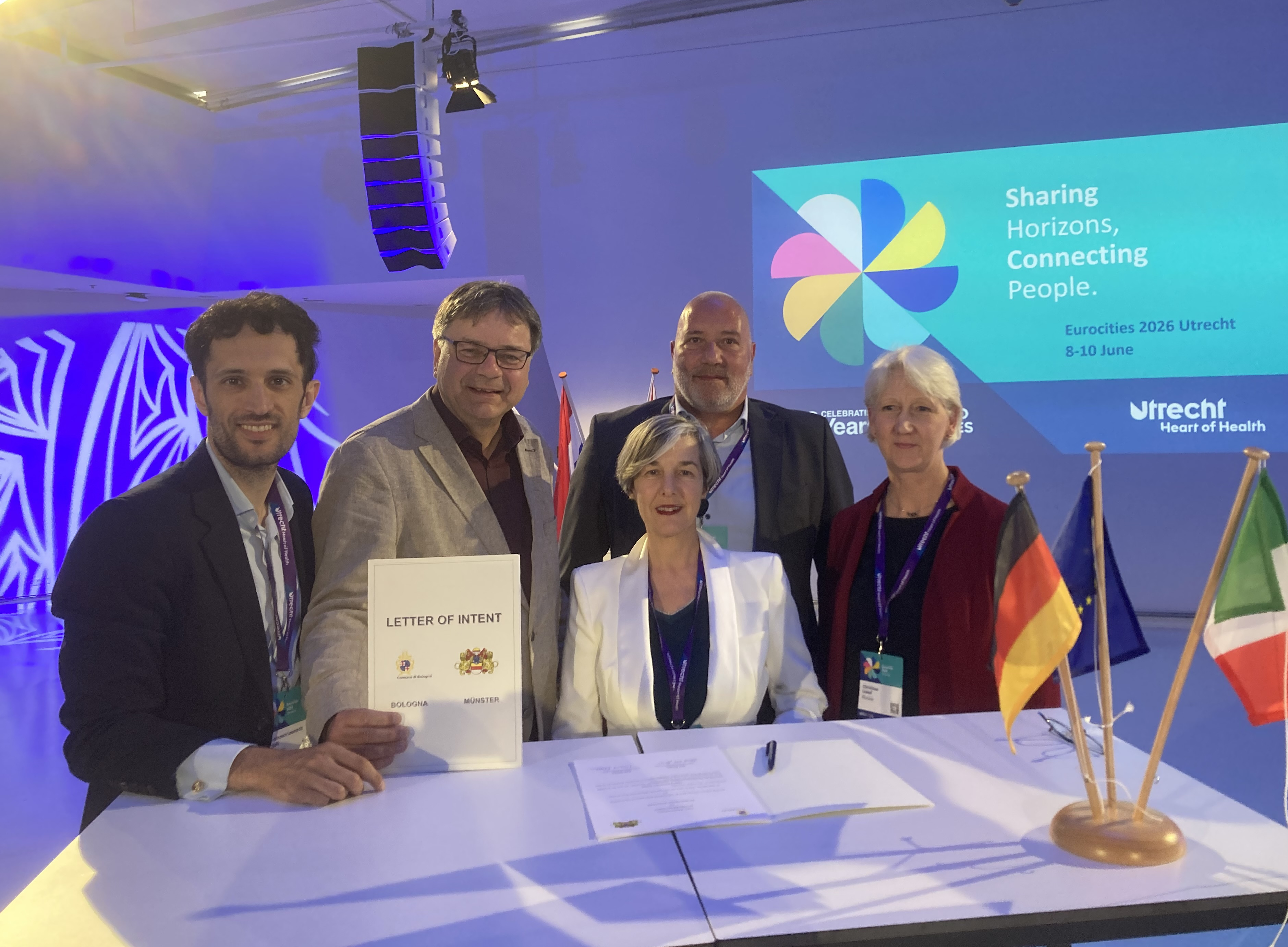 Gruppenfoto: Fünf Personen stehen vor dem Tisch und lächeln in die Kamera. Ein Mann hält eine Urkunde in der Hand. Auf dem Tisch stehen kleine Länderfähnchen.