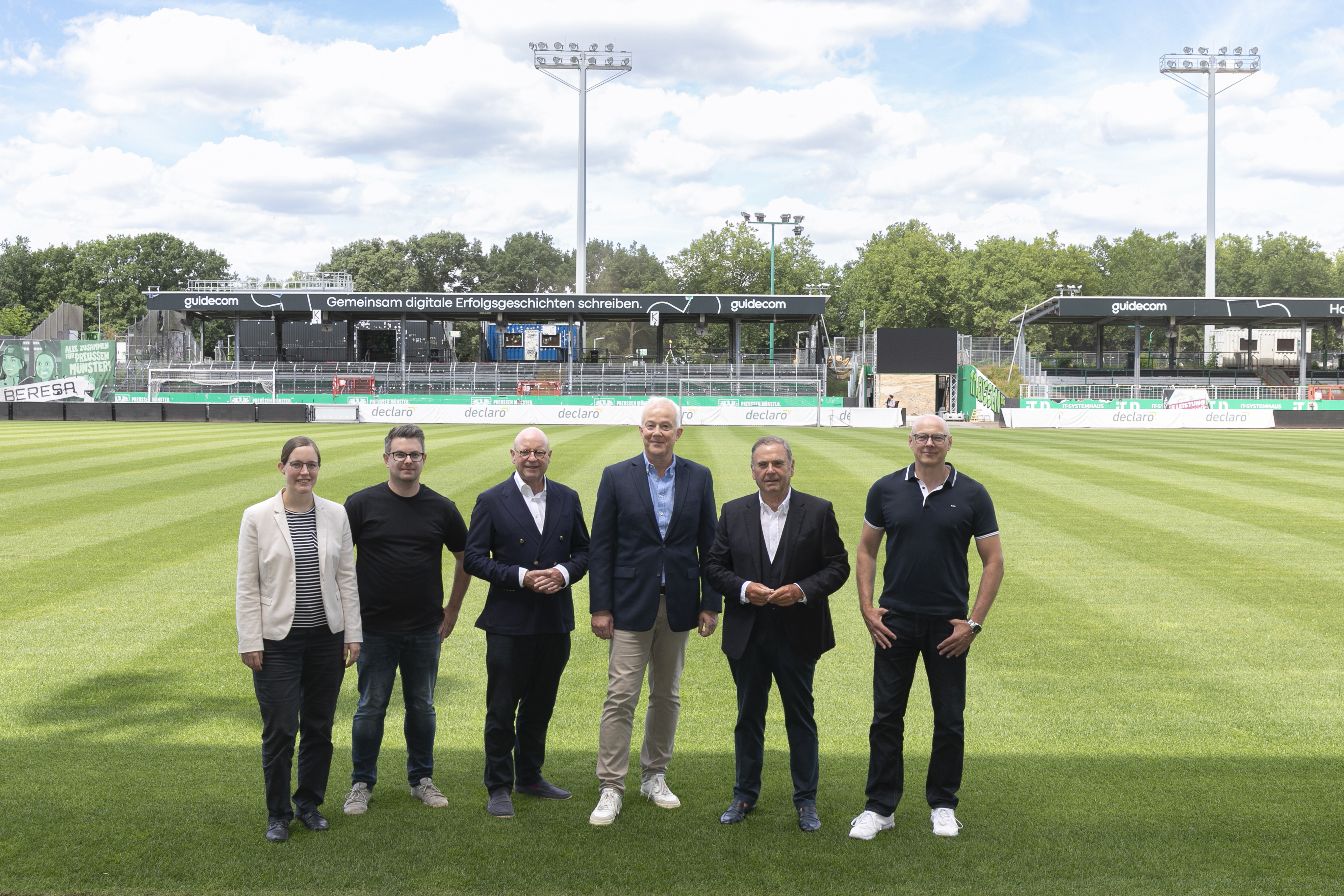 Die sechs Personen stehen nebeneinander auf dem Spielfeld des Stadions; im Hintergrund ist die Gegengerade.