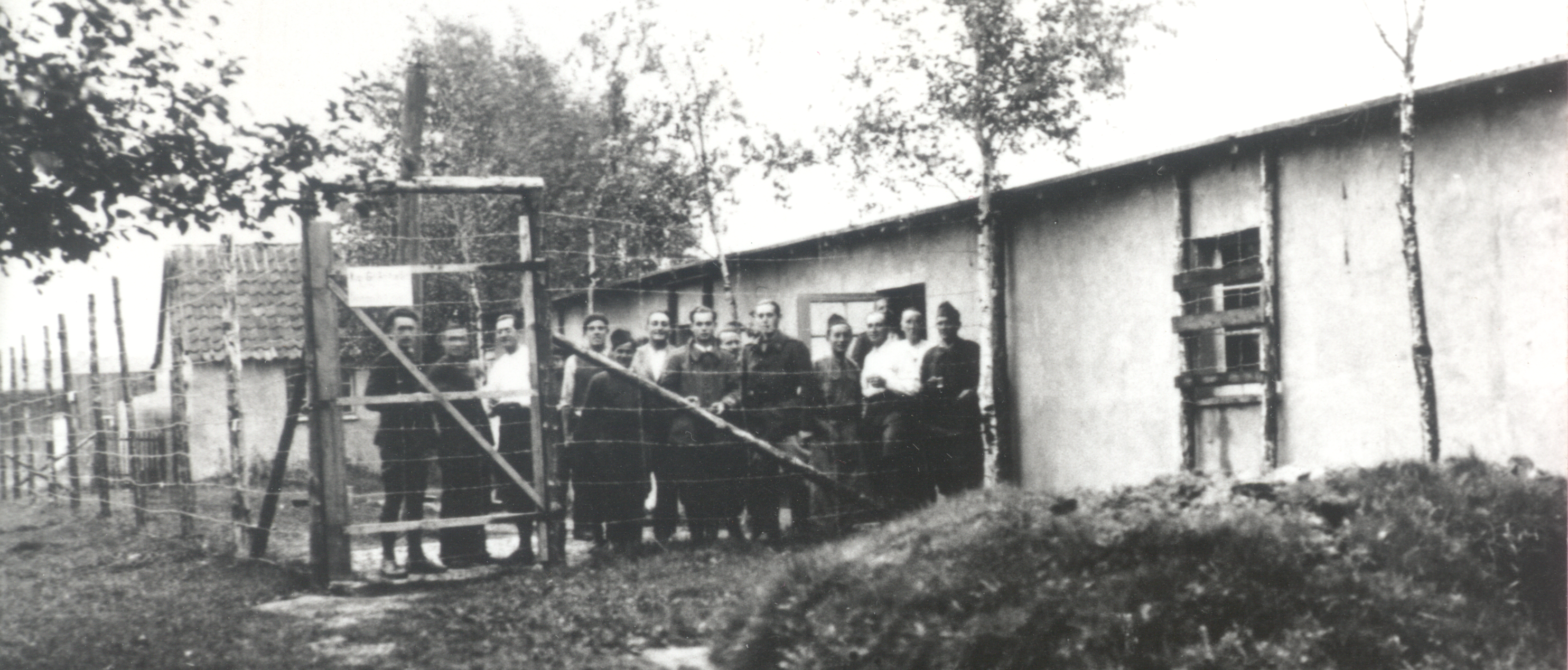 Mehrere Kriegsgefangene hinter einem Zaun eines Lagers blicken in die Kamera