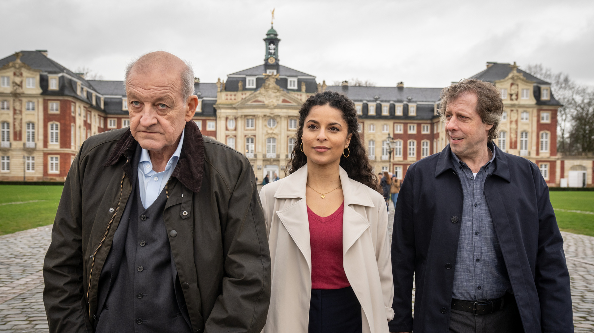 "Wilsberg - Über dem Gesetz": Wilsberg (Leonard Lansink), Dr. Tessa Tilker (Patricia Meeden) und Ekki Talkötter (Oliver Korittke) laufen über den Vorplatz eines Schlosses. Die Fassade des historischen Gebäudes erhebt sich im Hintergrund.