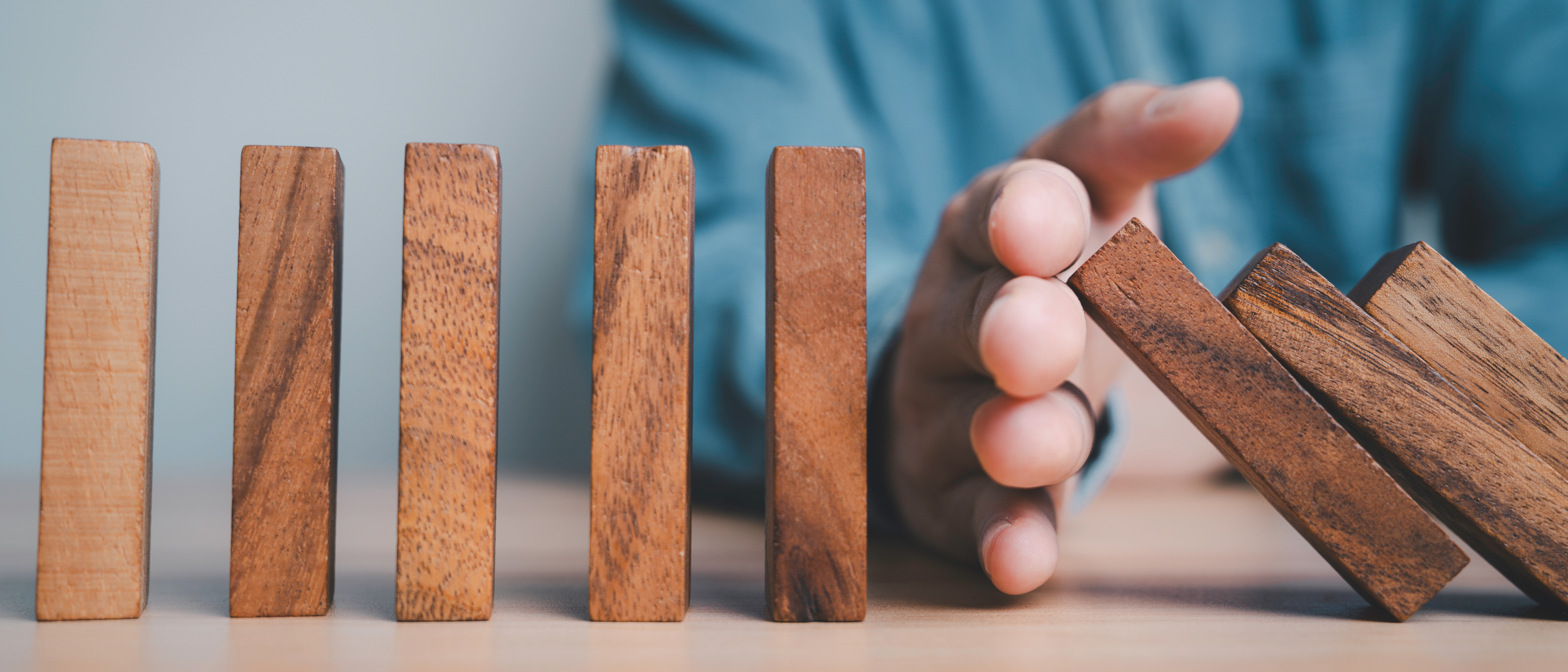 Dominosteine stehen auf einem Tisch, einige sind bereits umgefallen. Eine Hand verhindert, dass alle umgestoßen werden