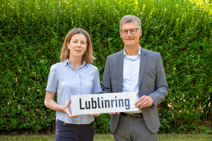 Eine Frau und ein Mann stehen vor einer grünen Hecke. Sie halten ein Schild mit der Aufschrift "Lublinring" in den Händen.
