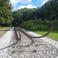 Auf dem Foto ist ein Denkmal zu sehen. Es besteht aus einer Eisenbahnschiene, deren vorderes Ende in Richtung Himmel gebogen ist, Eisenbahnschwellen aus Holz und weißen und grauen Steinen im Kiesbett der Schienen.