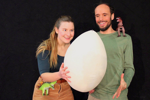 Eine Performerin hält ein riesiges Ei in Händen. Sie und ihr Schauspielkollege lachen beide mit Blick auf das gigantische Ei.