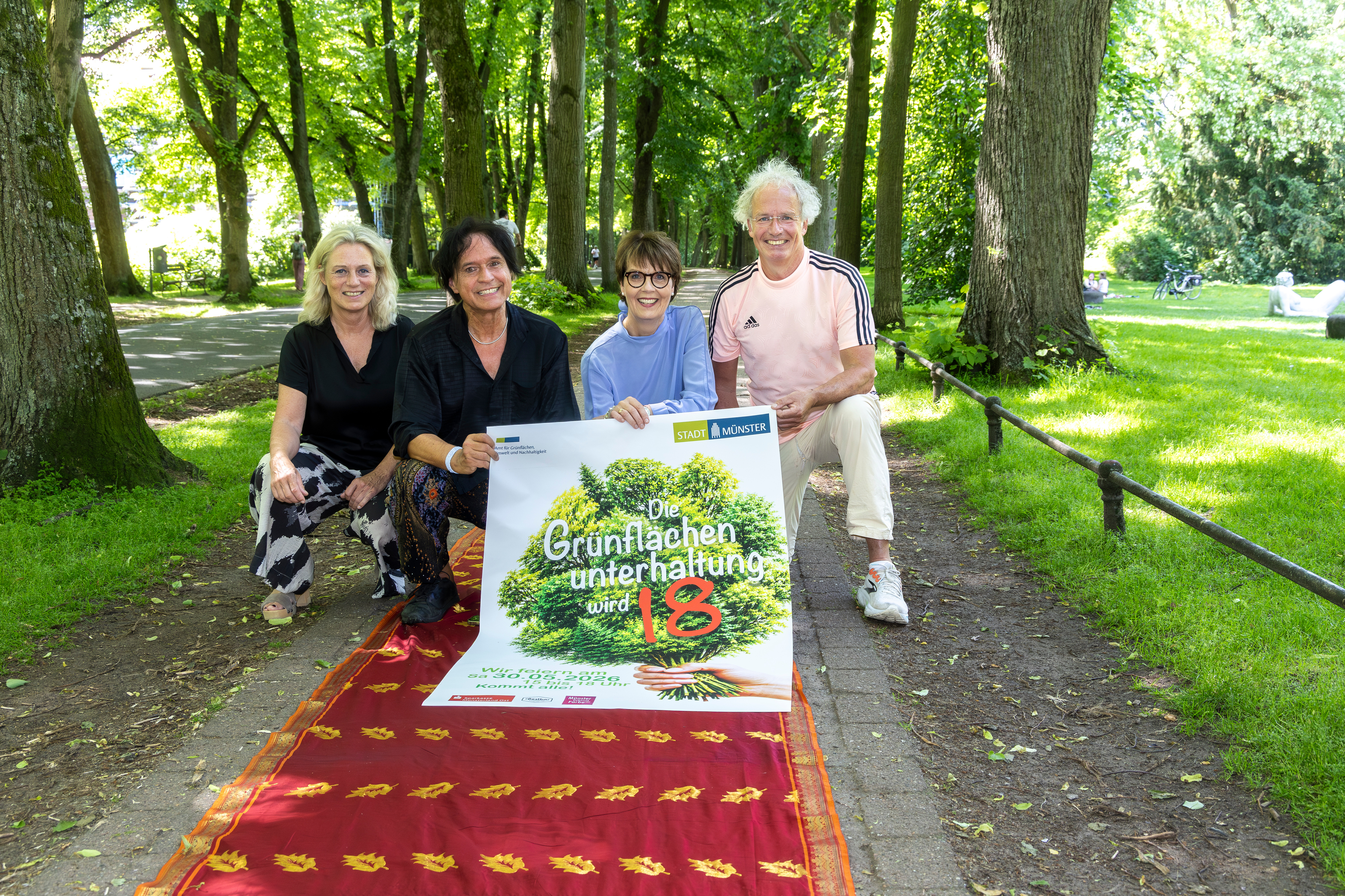 Vier Personen hocken nebeneinander und halten ein Plakat in der Hand, auf die Grünflächenunterhaltung beworben wird