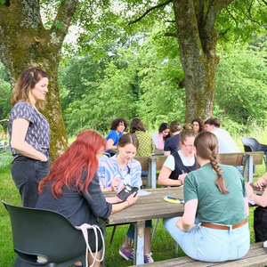 Eine Gruppe Frauen sitzt auf einer Wiese an einem Holztisch. Eine weitere Frau steht links neben dem Tisch.
