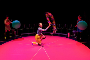 Eine Performerin kniet auf einem knallrosa Boden und schwingt ein Glitterband. Im Hintergrund steht jeweils rechts und links eine Person und hält Musikschlägel in der Hand.