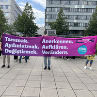 Drei Menschen demonstrieren mit einem Banner, auf dem 'Anerkennen, Aufklären, Verändern' steht.