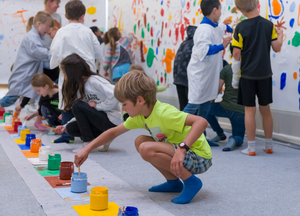 Eine Gruppe von Kindern malt gemeinsam an einer langen Leinwand. Im Vordergrund stehen Farbgläser.