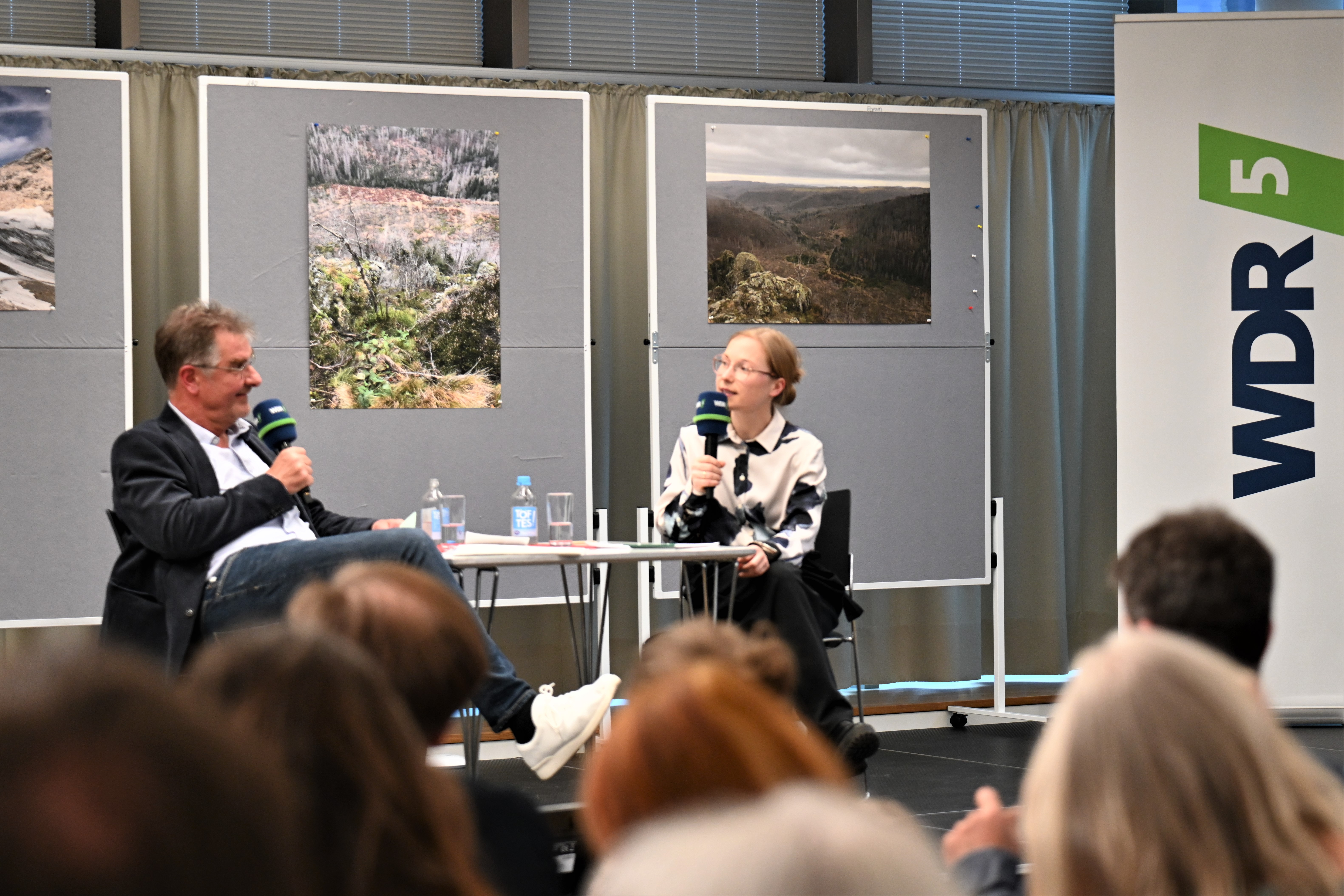 Moderator Jürgen Wiebicke, Mann in dunklem Jacket, und Dr. Lorina Buhr, junge blonde Frau sitzen auf einem Podium