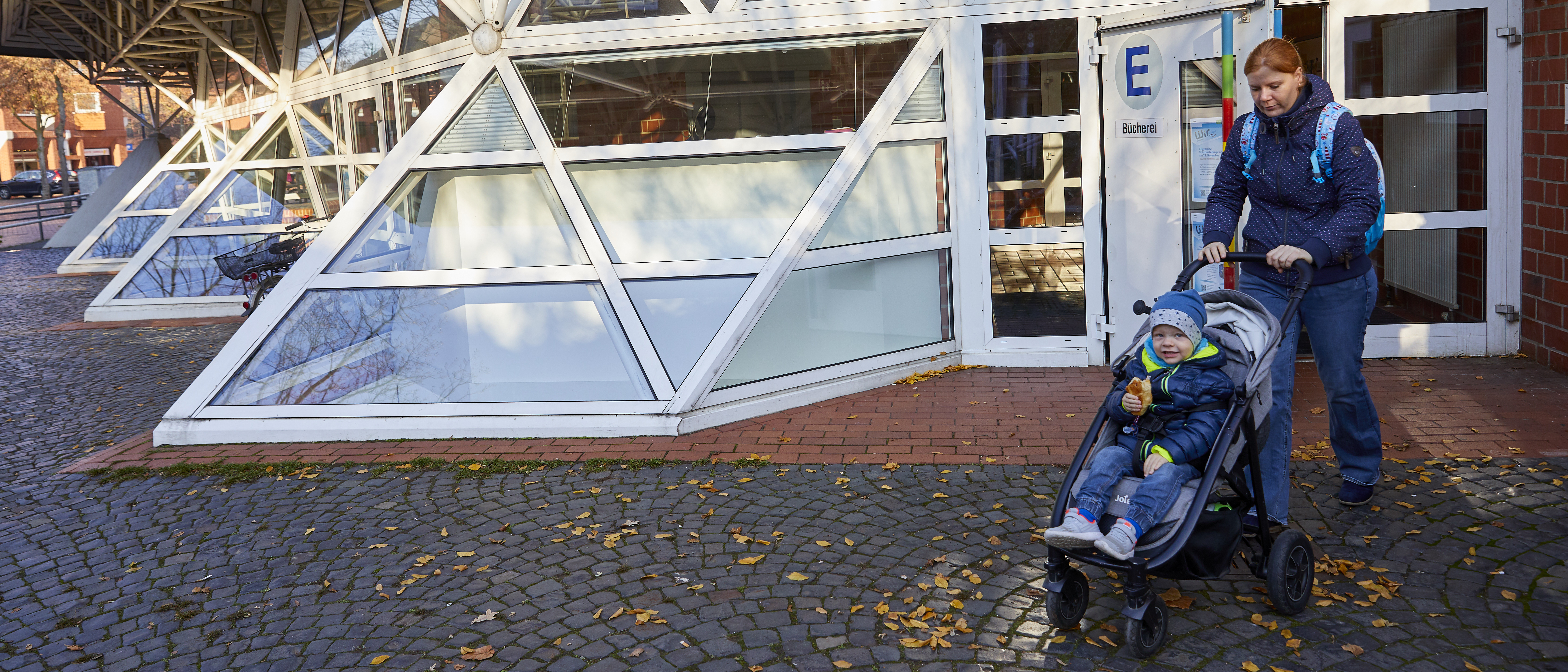 Außenaufnahme Bücherei Kinderhaus: Frau mit Kleinkind im Kinderwagen verlässt Gebäude
