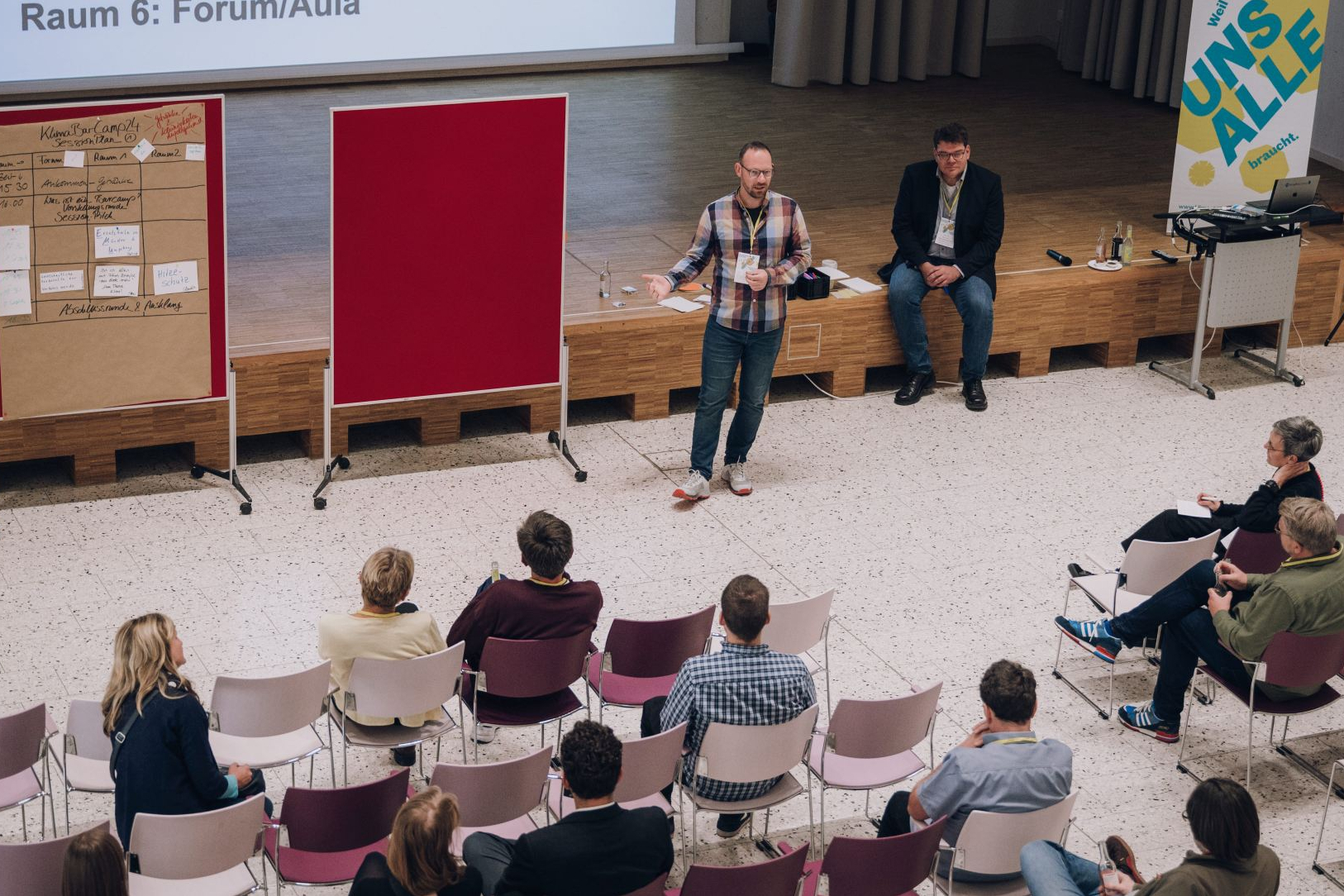 Blick auf Bühne und Zuhörer von schräg oben, während ein Teilnehmer des KlimaBarCamps 2024 einen Vortrag hält.