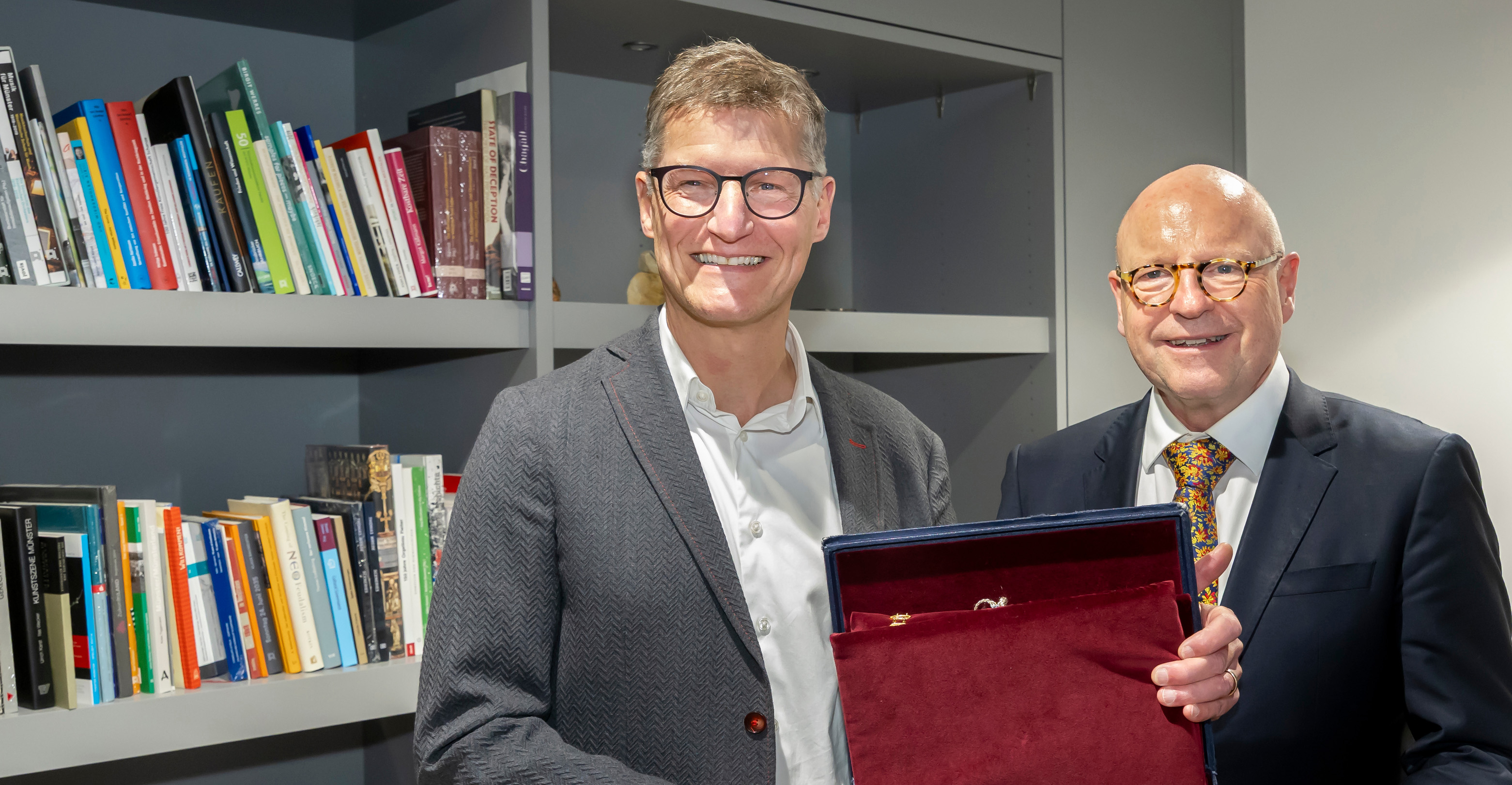 Zwei Männer lächeln in die Kamera und halten eine Schmuckschatulle mit einer großen Kette darin in den Händen