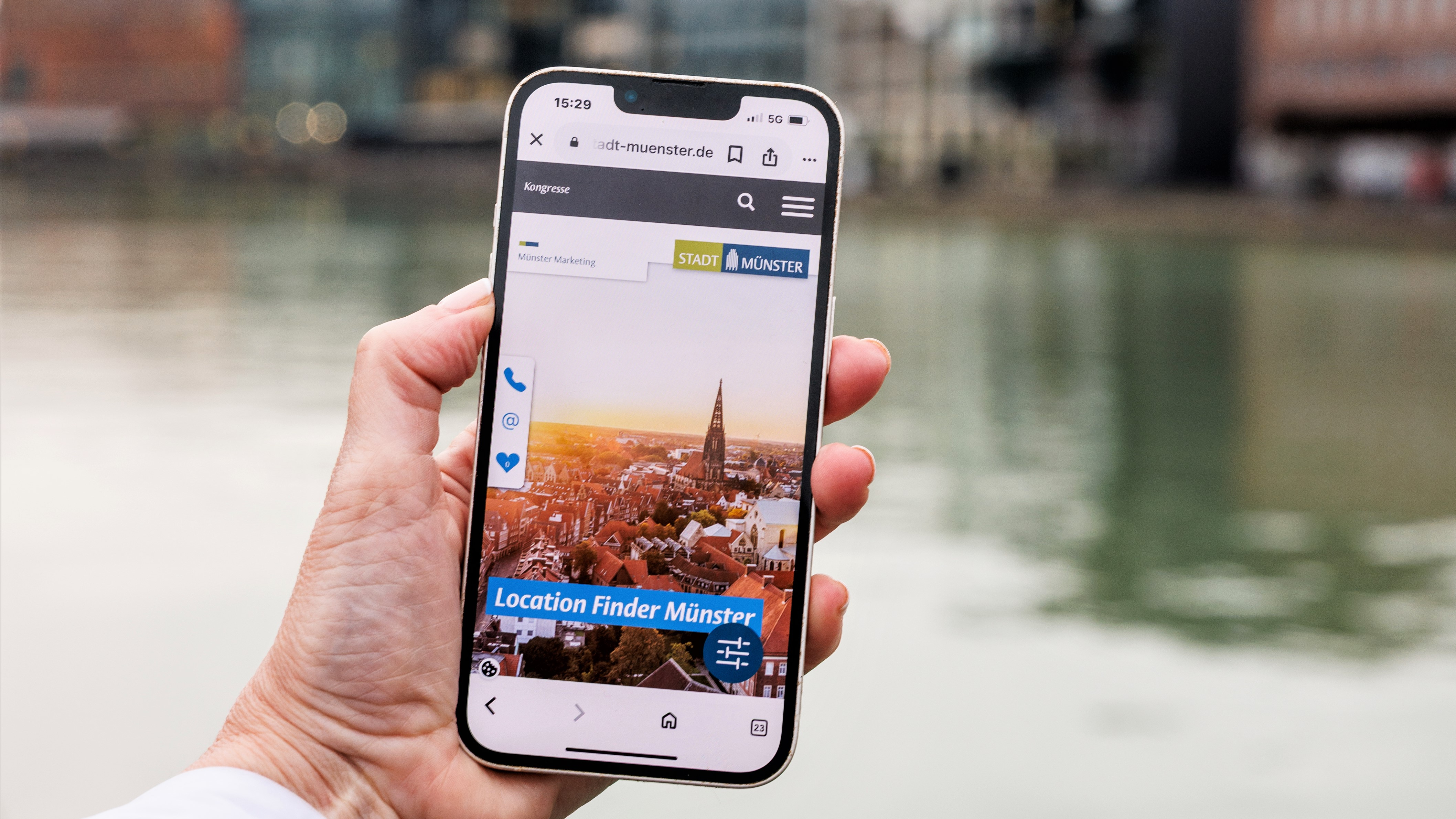 Eine Hand hält ein Smartphone, auf dem der Location Finder von Münster Marketing zu sehen ist, im Hintergrund liegt der Kreativkai in Münsters Hafen.