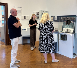 Zwei Frauen und ein Mann stehen vor einem weißen Regal. Am Regal ist eine durchsichtige Vitrine angebracht. In der Vitrine steht eine grüne Uniform.
