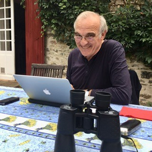 Fotografie zeigt den Autor Wilhelm Gruber sitzend am Tisch, auf dem ein aufgeklapptes Laptop und ein Fernglas steht.