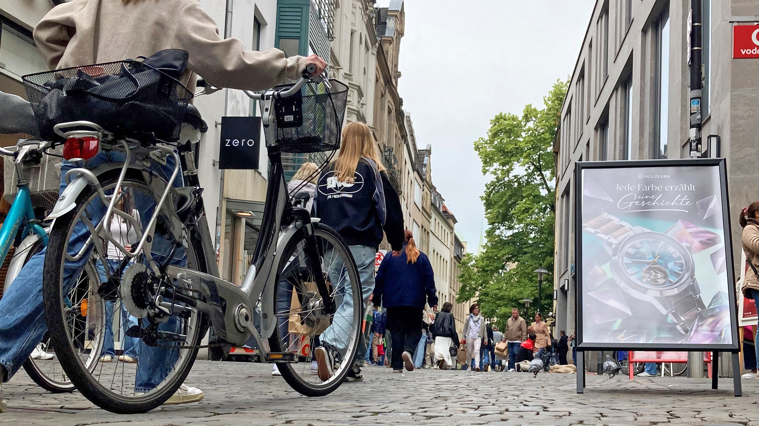 Fußgänger und eine Passantin, die ihr Fahrrad schiebt, in einer Einkaufsstraße mit Geschäften in der Innenstadt von Münster.