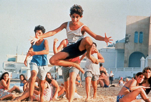 Ein Jugendlicher springt mit offenen Armen und angezogenen Beinen frontal Richtung Kamera. Er befindet sich auf einem Strand