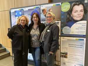 Gruppenfoto: drei Frauen stehen im Raum, im Hintergrund eine große Wand mit Plakaten